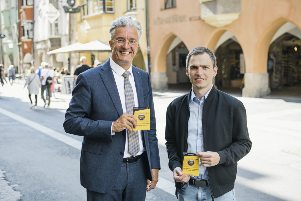 Fotografie von Patrick Pixner und Michael Perger in der Innsbrucker Innenstadt. Beide halten die Inn-Taler Gutscheinkarte in den Händen, welche als analoge und digitale Währung die regionale Kaufkraft fördert.